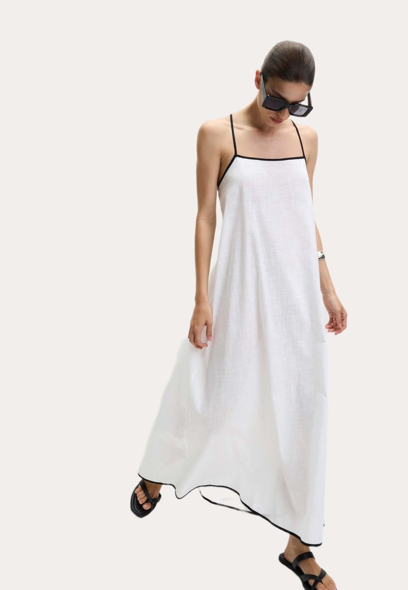 Robe maxi blanche fluide avec de fines bretelles noires et un contraste de bordure noire le long de l'ourlet. Associée à des sandales plates noires, mettant en avant une coupe décontractée.
