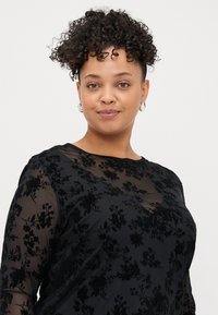 Haut noir à manches longues avec un motif floral transparent. Caractérisé par un col rond et une texture douce, avec une silhouette ajustée tout au long.