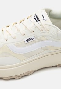 Cream Vans sneakers feature a textured upper with a white swoosh, beige paneling, and a thick sole. Laces are light beige.