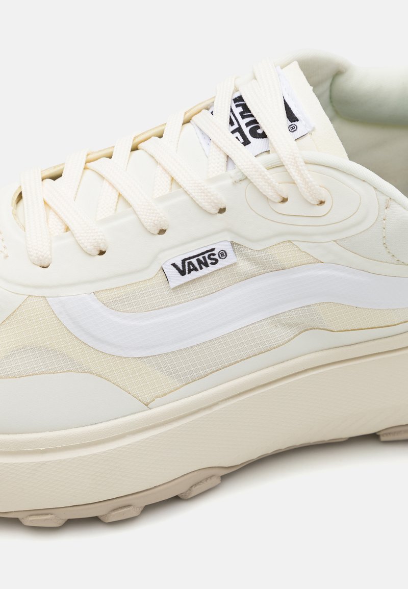 Las zapatillas Vans de color crema tienen una parte superior texturizada con un swoosh blanco, paneles beige y una suela gruesa. Los cordones son de un beige claro.