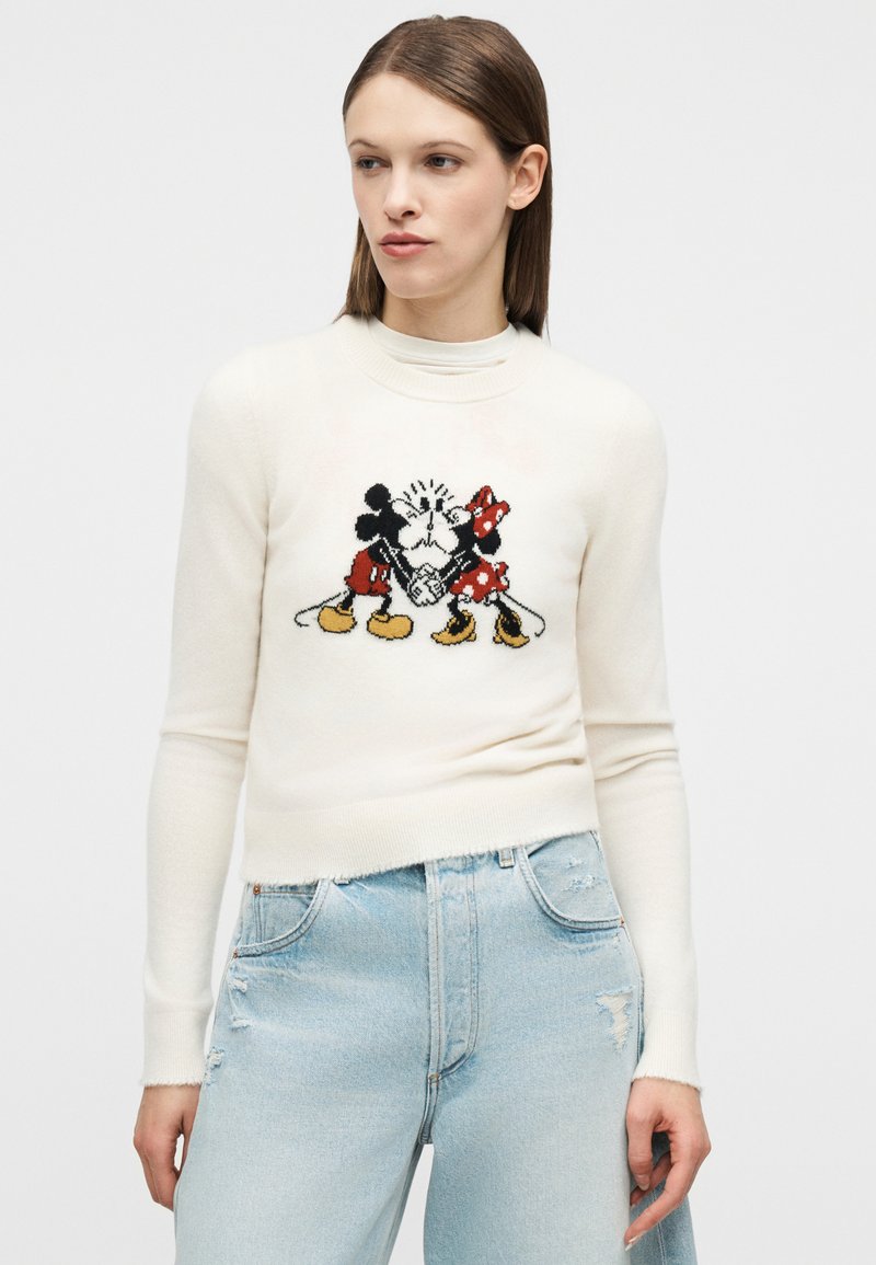 Frau trägt einen cremefarbenen Pullover mit gestricktem Mickey- und Minnie-Maus, die Händchen halten, kombiniert mit hellblauen, zerrissenen Jeans.