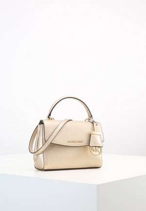 Mała złota torebka Michael Kors z górnym uchwytem, długim paskiem i zawieszką z logo MK na białej powierzchni i tle.