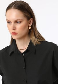 Black button-up shirt with a collar, featuring a smooth texture. Gold hoop earrings with a shiny finish, complementing a thin gold chain necklace.