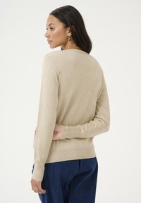 Beige kötött pulóver hosszú ujjal, bordázott mandzsettával és szegéllyel, testhez simuló fazonnal és finom textúrával. Kék farmernadrággal viselve.