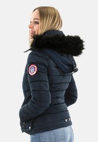 Mujer con una chaqueta acolchada azul marino y capucha de piel negra, con un parche redondo rojo y blanco en el brazo izquierdo, con las manos en los bolsillos.