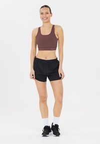 Sportbeha in donkerrood, gecombineerd met zwarte shorts. Glad materiaal, sportieve snit, elastische tailleband en zwarte sneakers met witte sokken.