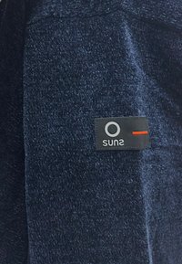 Tessuto blu navy con una superficie testurizzata, caratterizzato da un'etichetta nera con il logo "sun2" e una linea di accento rossa nella parte inferiore.