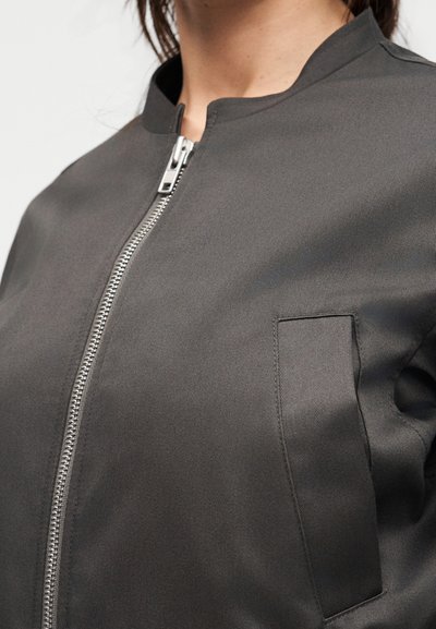 Κοντινό πλάνο ενός ατόμου που φοράει σκούρο γκρι μπουφάν με φερμουάρ, όρθιο γιακά και ορθογώνια τσέπη στο στήθος στην αριστερή πλευρά.