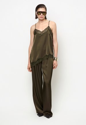 Vrouw draagt olijfgroene satijnen top met kanten rand, bijpassende wijde broek, grote getinte zonnebril en een gouden manchet armband.