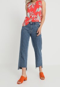 Top sin mangas rojo con flores y patrones de hojas blancas, combinado con jeans de pierna ancha azul de cintura media y zuecos de tacón bloque naranja.