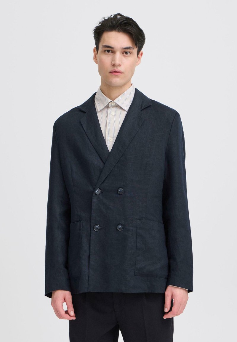 Jeune homme portant un blazer sombre à double boutonnage sur une chemise claire rayée à boutons, debout devant un fond clair uni.