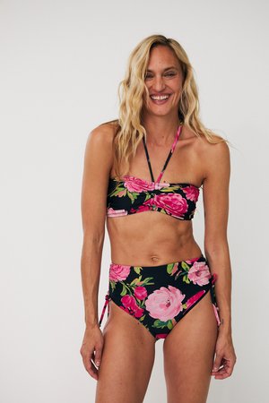 Smiling woman with wavy blonde hair wearing a black bikini with large pink and green floral prints, standing against a white background.