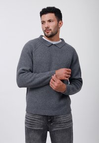 Grå ribbad tröja med krage, som har långa ärmar och en avslappnad passform. Texturen är tjockstickad, i kombination med mörkgrå jeans.