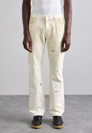 White denim jeans featuring distressed stains, a straight-fit design, and five-pocket styling. Paired with black shoes and a fitted white tank top.