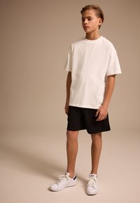 White cotton t-shirt with short sleeves, paired with black shorts and white sneakers with blue accents, positioned against a neutral background.