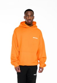 Sudadera con capucha naranja de material suave, con un bolsillo delantero y el logo blanco "MULTIPLY". Combinada con pantalones de chándal negros que presentan el mismo logo de la marca.