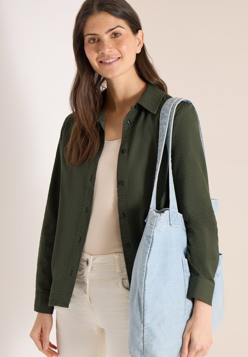 Ung kvinde iført mørkegrøn skjorte med knapper og cremefarvede bukser, bærende en stor lyseblå tote-taske på skulderen, smilende.