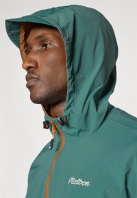 Φόρμα με κουκούλα σε σκούρο μπλε ύφασμα με λεία υφή. Διαθέτει καφέ εσωτερικό, μπροστινό φερμουάρ με πορτοκαλί λεπτομέρειες και κεντημένο λογότυπο.