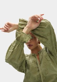 Lichtgroene blouse met opgezette mouwen, voorzien van bloemige borduursels en een gestrikt decolleté. De stof heeft een gestructureerde afwerking en valt soepel.