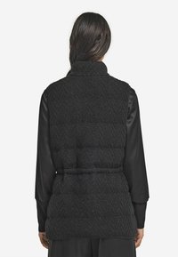 Svart vadderad väst med ett fiskbensmönster, hög krage och midjebälte. Tyget har en subtil glans och en strukturerad yta.