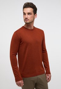 Maglione a maniche lunghe color ruggine con scollo rotondo, texture morbida e polsini a costine. Indossato con pantaloni beige, su uno sfondo neutro.