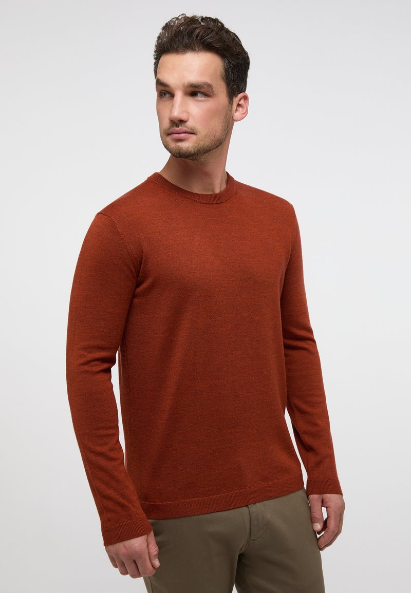 Maglione a maniche lunghe color ruggine con scollo rotondo, texture morbida e polsini a costine. Indossato con pantaloni beige, su uno sfondo neutro.