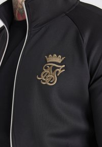 Giacca nera con zip, caratterizzata da un logo ricamato in oro e dettaglio a corona, realizzata in un tessuto liscio ed elastico con un design aderente.