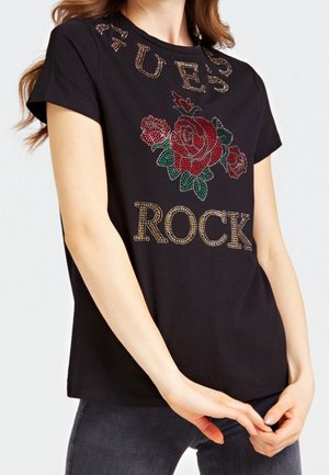 Kobieta nosząca czarną koszulkę z różami z kryształków i napisem "GUESS ROCK" z przodu, w połączeniu z ciemnymi jeansami.