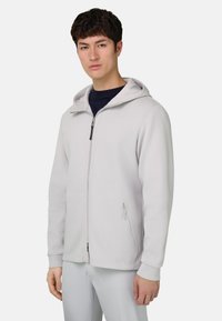 Sweat à capuche gris clair avec fermeture éclair, une capuche, deux poches latérales, des poignets côtelés et une texture lisse. Porté par-dessus un t-shirt foncé.