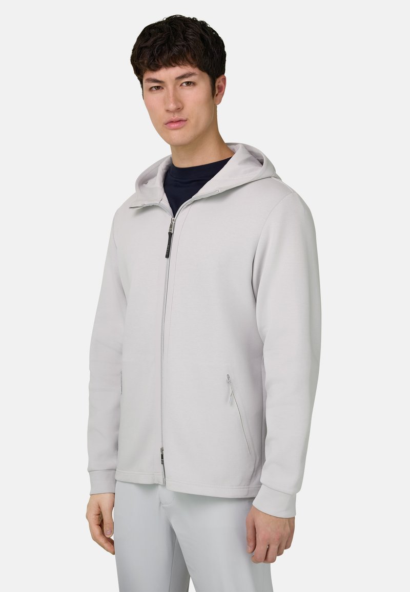 Sweat à capuche gris clair avec fermeture éclair, une capuche, deux poches latérales, des poignets côtelés et une texture lisse. Porté par-dessus un t-shirt foncé.