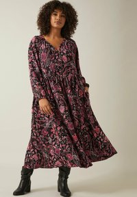 Robe longue noire à motifs floraux roses et blancs, décolleté en V, manches longues et taille froncée, associée à des bottes noires montantes.