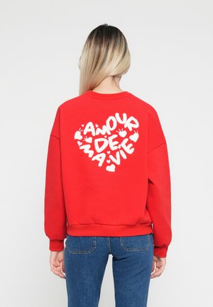 Rød hettegenser laget av bomull, med et fluffy hvitt hjertegrafikk og teksten "AMOUR DE MA VIE" på baksiden. Standard passform med ribbestrikkede mansjetter.