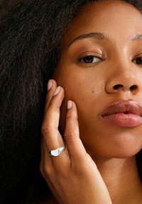 Srebrny pierścionek z subtelnym kształtem serca, noszony na dłoni z naturalnym, jasnym lakierem do paznokci; w tle widoczne teksturowane ciemne włosy i skóra twarzy.