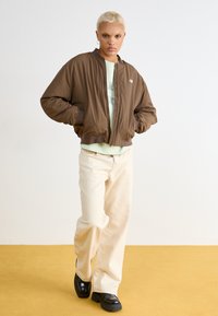 Jaqueta bomber leve em tons de castanho com punhos canelados, fecho de correr e bolsos laterais, usada sobre uma blusa verde menta e combinada com calças largas em tom creme.