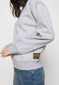 Felpa grigia con scollo tondo, maniche lunghe e polsini a coste. Presenta un patch logo nero e giallo sul lato sinistro.