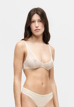 Femme aux longs cheveux bruns portant un soutien-gorge transparent beige clair et des sous-vêtements assortis sur un fond gris clair uni.