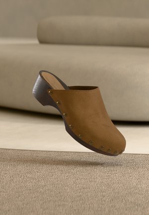 Sabot en daim marron avec talon en bois et clous en laiton, flottant au-dessus d'un tapis beige dans une pièce aux tons neutres.