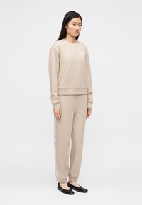 Μπεζ φούτερ και ταιριαστός αθλητικός παντελόνι από μαλακό ύφασμα, με λογότυπο στο στήθος και κάθετη σήμανση κατά μήκος του παντελονιού. Μαύρα φλατ παπούτσια.