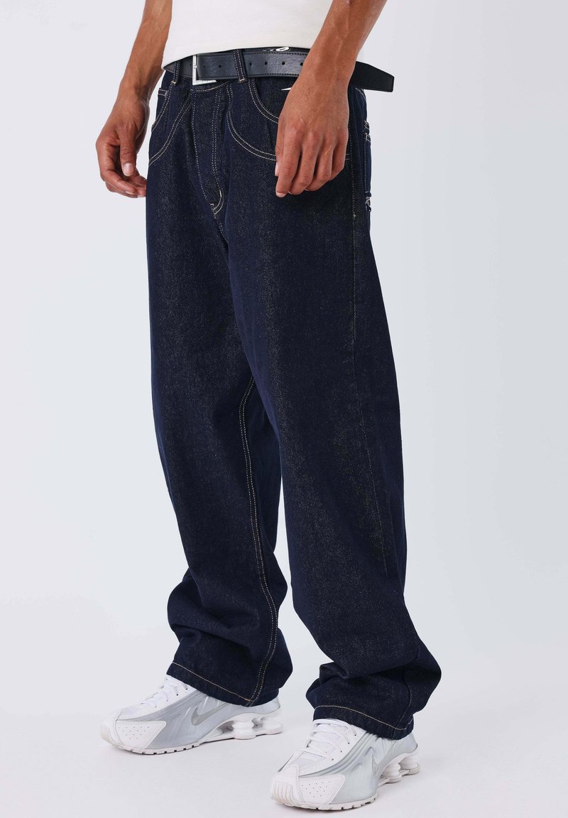 Vaqueros de mezclilla azul oscuro con un corte holgado, que presentan costuras amarillas, bolsillos delanteros y un cinturón negro. Combinados con zapatillas deportivas blancas.