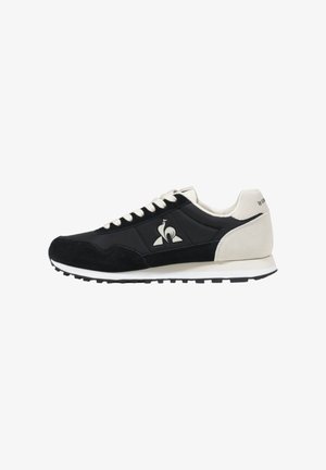 Zapatilla negra con parte superior de ante y cuero, suela de goma blanca, detalles en color crema y un logotipo en el lateral. Estilo de cordones con punta redondeada.