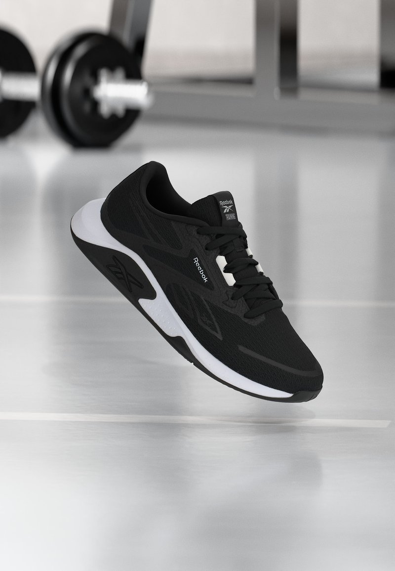 Μαύρο και άσπρο αθλητικό παπούτσι Reebok που αιωρείται σε γυμναστήριο με θολωμένα βάρη μπάρα στο φόντο, σε γυαλιστερό δάπεδο.