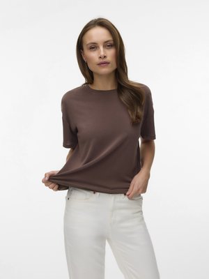 Γυναίκα με μακριά καστανά μαλλιά που φοράει καφέ κοντομάνικη μπλούζα και λευκό παντελόνι, κρατώντας το τελείωμα της μπλούζας με το ένα χέρι.