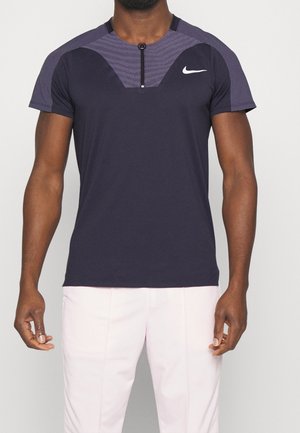 Homme portant un T-shirt de sport bleu marine foncé avec une fermeture éclair à l'avant et un logo Nike blanc, associé à un pantalon rose clair.