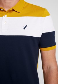 Homme portant un polo à manches courtes avec des blocs de couleur jaune moutarde, blanc et bleu marine, et un petit logo brodé en forme d'oiseau.