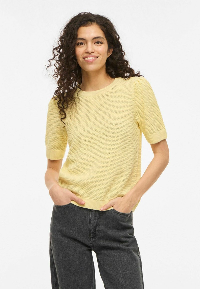 Femme souriante aux cheveux bouclés portant un haut en maille jaune à manches courtes et un jean taille haute foncé, mains dans les poches, sur un fond uni.