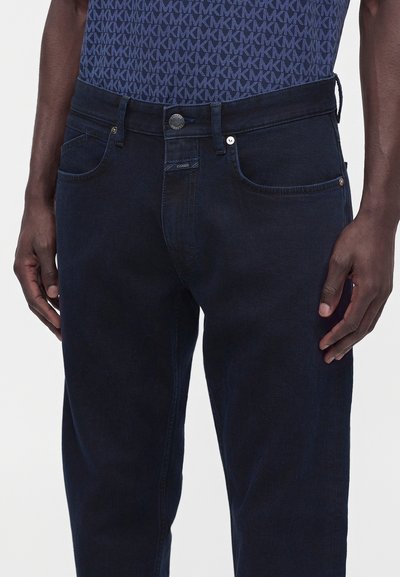 Jean bleu foncé porté par une personne, les mains détendues le long du corps, associé à une chemise bleue avec un motif répétitif "MK".