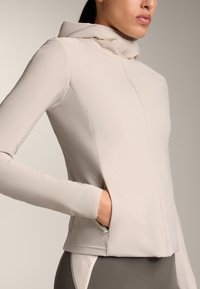 Ženska, oblečena v svetlo bež jopico z zadrgo in kapuco, z roko v stranskem žepu, prikazana od ramen do bokov na nevtralnem ozadju.