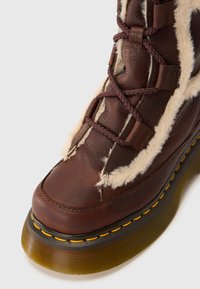 Brun læderstøvle med faux pelskant, rund tå og dekorativ gul syning langs sålen. Snøredetaljer har tykke brune snørebånd.