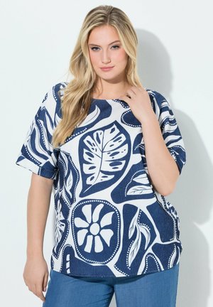 Blonde woman wearing a blue and white patterned short-sleeve top and blue pants, standing against a plain white wall.