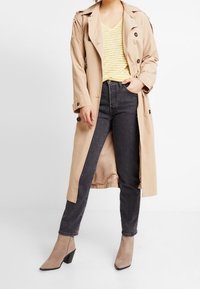 Beige trenchcoat met dubbelzijdige knopen, gelaagd over een geel en wit gestreept t-shirt, gecombineerd met donkere jeans en taupe enkellaarsjes.